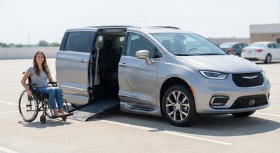 A woman in a wheelchair smiling next to a silver Chrysler Pacifica minivan with its wheelchair ramp deployed in a parking lot.
