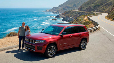 Couple enjoying a scenic coastal view next to a red Jeep SUV rental car on a USA road trip adventure.