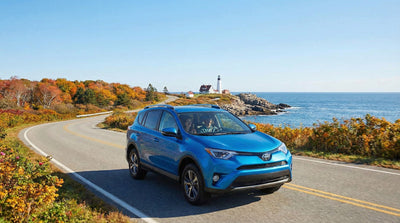 Happy couple driving a blue Toyota Rav4 car rental along a scenic coastal road towards Acadia National Park.