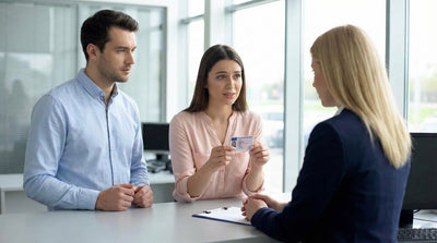 A couple at a car rental desk; the woman presents her driving licence while the man next to her has empty hands and a concerned look.