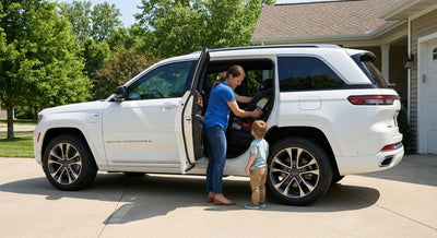 A mother installing a child car seat into the back of a white Jeep Grand Cherokee L while her young son watches, in a suburban driveway.