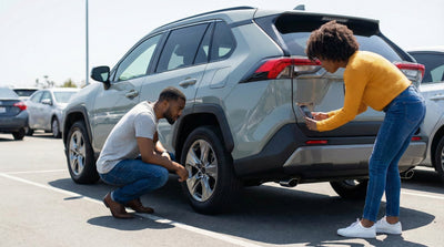 A couple carefully inspects the tires and body of a grey rental car in a sunny parking lot before picking it up.