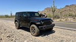 A black Jeep Wrangler rental SUV parked on a gravel shoulder next to a desert highway, with a person examining the windshield under bright desert sun.