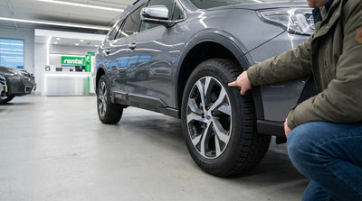 A person kneels in the snow to inspect the tires on a car rental on a New York street