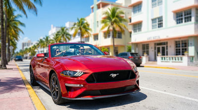 A blue convertible car hire drives down a sunny street lined with palm trees and Art Deco buildings in Miami
