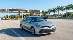 Driver in a white car rental approaching a sunlit highway toll booth in Florida