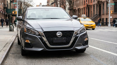 A gray car hire driving on a New York street with its front bumper and missing number plate visible