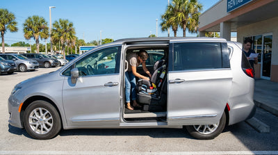 A parent installs a child car seat into the back of a white SUV at a car hire location in sunny Florida