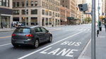 A car rental driving on a busy New York street with a clearly marked red bus lane and traffic camera overhead