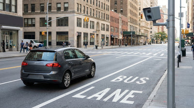 A car rental driving on a busy New York street with a clearly marked red bus lane and traffic camera overhead