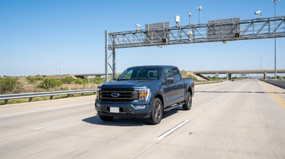 A car rental driving on a Texas highway toward a large electronic toll gantry under a clear blue sky