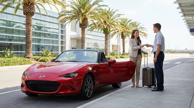 A traveler with luggage waits for a car rental at a sunny Miami airport curb lined with palm trees