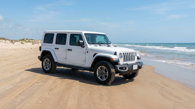 A white car rental SUV drives on the sand at South Padre Island, Texas, with blue ocean waves along the shore