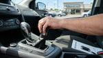 A driver's hand on the stick shift of a car rental with a scenic United States highway seen through the windshield