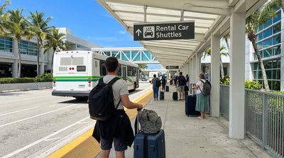 Travelers wait under the purple car rental shuttle sign outside LAX Terminal 1 in Los Angeles