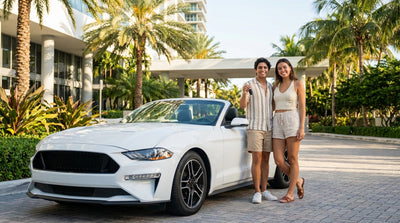 A young person smiling next to their red convertible car rental on a sunny street in Miami