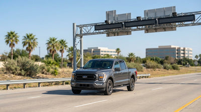 A silver car rental driving under an electronic toll gantry on a wide Texas highway