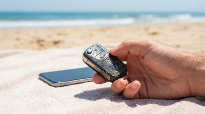 A hand holding a wet key fob for a car rental on a sunny Florida beach with the ocean in the background