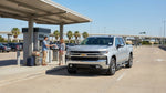 A silver car rental drives under a large electronic toll sign on a sunny multi-lane highway in Texas