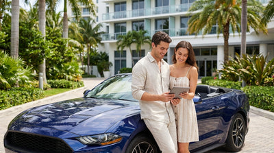 A young driver smiling next to a red convertible car rental on a sunny street with palm trees in Miami