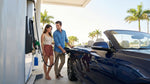 A person refueling their white car rental at a gas station pump under a big, sunny Texas sky