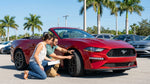 A person checking the tires of a white car rental sedan parked under palm trees on a sunny Florida street