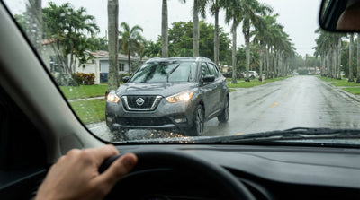 A car rental drives with headlights on through a heavy rainstorm on a palm tree-lined street in Miami