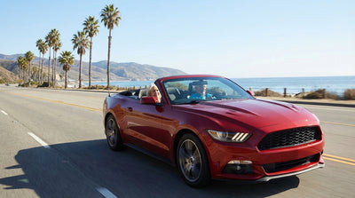 A senior man smiles next to his convertible car hire on a sunny coastal road in California