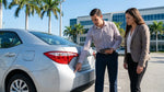 Person inspecting a scratched bumper on a car rental parked along a sunny beach road in Florida