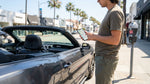A driver using a mobile phone next to their car rental on a palm tree-lined street in Los Angeles