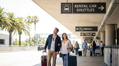 Travelers with luggage wait for a car rental shuttle under a purple sign at LAX in Los Angeles