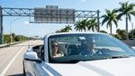 A car rental drives under a toll-by-plate gantry on a sunny, palm-lined highway in Florida