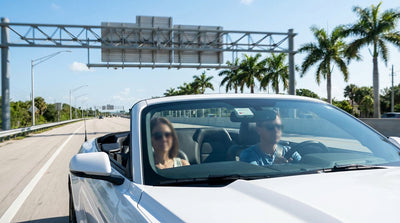 A car rental drives under a toll-by-plate gantry on a sunny, palm-lined highway in Florida