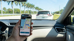 A person in a car rental uses a smartphone with a map app while driving on a sunny Florida road with palm trees