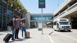 Travelers wait for a car hire shuttle bus under a purple sign outside LAX Terminal 2 in Los Angeles