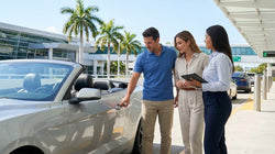 Person inspecting a scratch on the bumper of a white car rental parked near palm trees in Miami