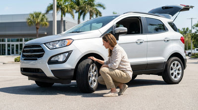 A person kneels to check the tire of their silver car rental on a sunny road in Florida