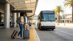 Travelers with luggage wait for a car hire shuttle bus at a sunny arrivals curb in Miami