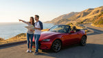 A convertible car rental driving down an open highway along the California coast at sunset