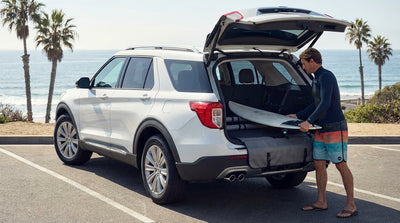 A surfer straps a surfboard to the roof of a red car hire parked on a sunny California coastal highway