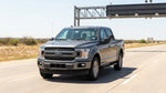 A modern car rental drives under a large electronic toll sign on a sunny multi-lane highway in Texas