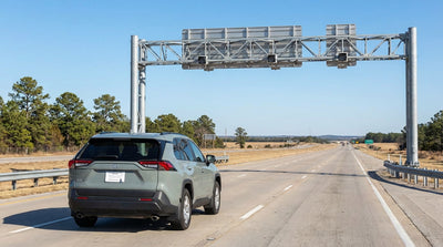 A modern car hire vehicle driving under a large electronic toll sign on a sunny Texas freeway