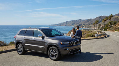 A Jeep Grand Cherokee car rental parked on a cliff overlooking the sunny Big Sur coast in California
