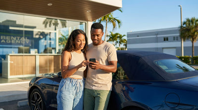 A white convertible car rental drives down a sunny Ocean Drive in Miami past colorful art deco hotels