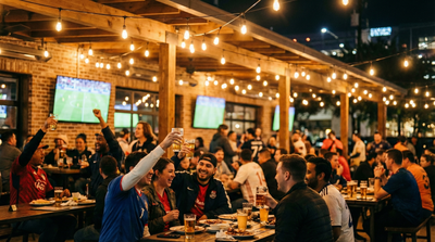 Fans cheering with beers at a packed outdoor football bar in Houston’s EaDo, watching a match on big screens under string lights. FIFA World Cup 2026.