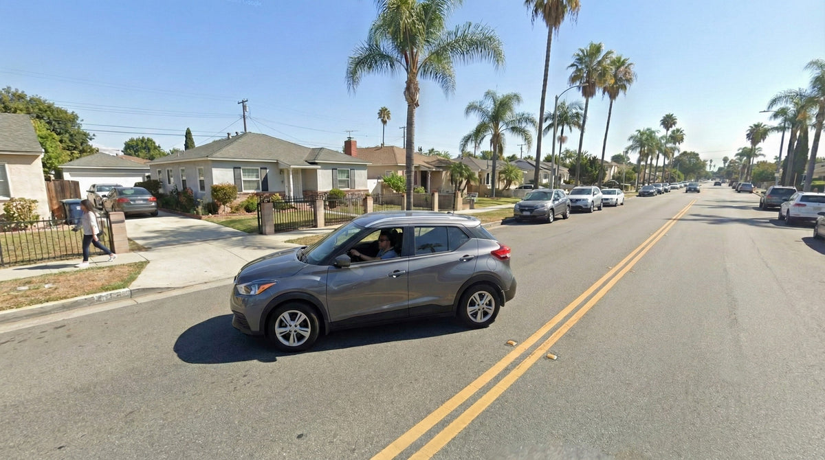 A grey Nissan Kicks drives down a sunlit residential street lined with palm trees and parked cars.