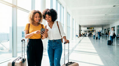 Two women with luggage checking car rental information on a phone at Miami Airport.