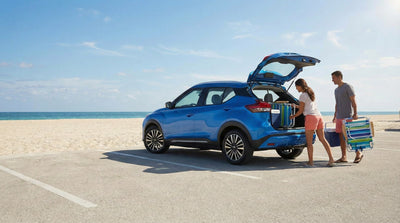 A blue Nissan Kicks SUV parked near a clean Miami beach, with a couple carefully loading beach gear into the trunk.