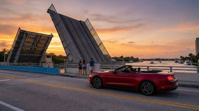 A red Ford Mustang Convertible rental car is stopped at a drawbridge rising at sunset in Miami, with people watching from the side.