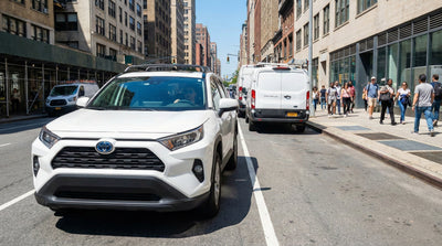 A white Toyota RAV4 rental car is shown navigating around double-parked vans on a busy city street in New York, with pedestrians on the sidewalk.